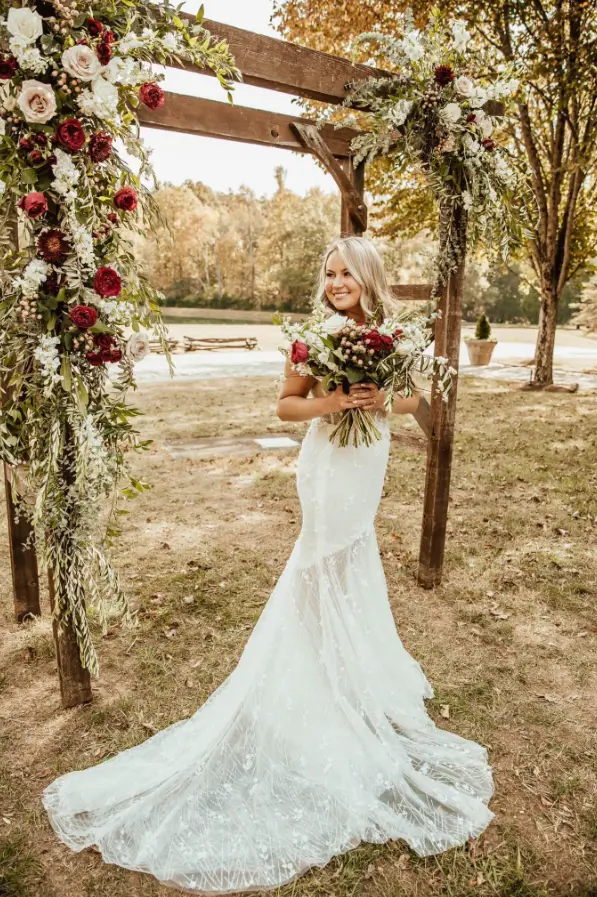 Bride holding a bouquet of deep red and cream roses beneath a wooden ceremony arbor